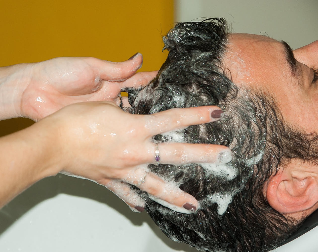 Mani di una persona che massaggiano delicatamente il cuoio capelluto con le dita, visti dall'alto, con capelli bagnati e gocce d'acqua che brillano