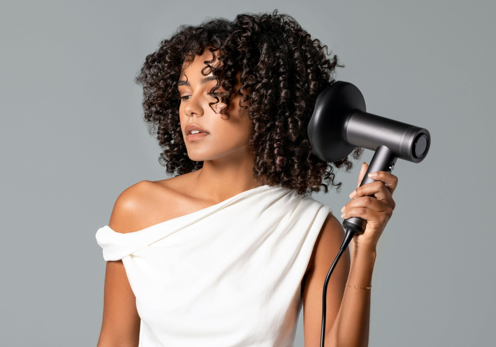 Donna con capelli castani crespi mentre asciuga i capelli con phon a temperatura moderata, utilizzando spazzola rotonda e prodotto anti-crespo
